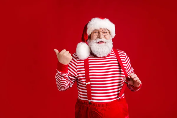 Photo of retired old man grey beard play straps direct thumb empty suggest noel sale space wear santa costume suspenders spectacles striped shirt headwear isolated red color background — Stock Photo, Image