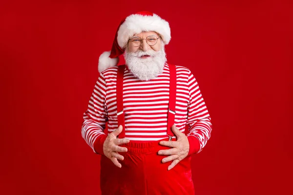 Zdjęcie emerytowanego dziadka szarej brody ręce trzymać duży brzuch paunch po uczęszczaniu fast food cafe nosić Santa stroje szelki okulary pasiasta koszula nakrycia głowy odizolowany czerwony kolor tła — Zdjęcie stockowe