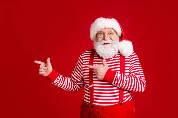 Photo de retraité vieux homme barbe blanche doigt direct espace vide suggérer solution intéressante porter Santa x-mas costume bretelles lunettes rayé chapeau chemise isolé couleur rouge fond — Photo