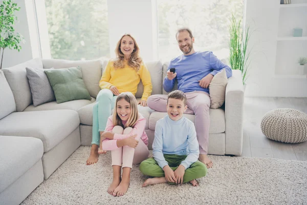 Full length photo of comfort family watch tv hold remote control two small kids sit floor mom tata na kanapie w domu — Zdjęcie stockowe