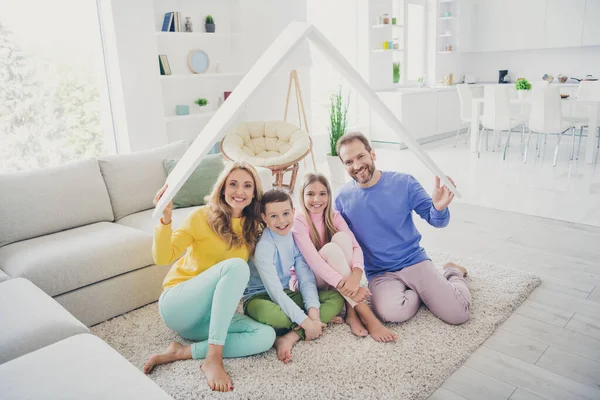Фотографія повнорозмірної гармонії сім'ї сидить на підлозі мама тато тримає паперовий дах над двома маленькими дітьми хлопчик-дівчинка в будинку в приміщенні — стокове фото