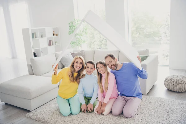 Full body photo of family mommy daddy sit floor carpet hold paper card roof above little kids boy girl in house indoors