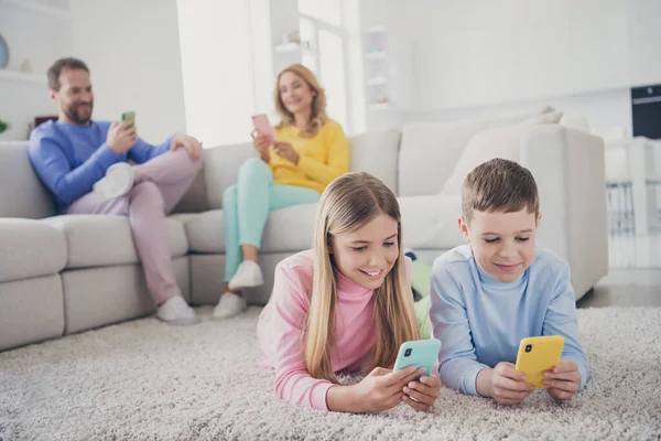 Foto de larga duración de la gente de la familia mamá papá sentarse sofá dos niños pequeños que mienten los mensajes de texto de la alfombra en el teléfono inteligente en casa en el interior —  Fotos de Stock