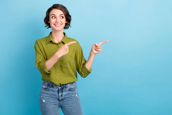 Foto der hübschen Dame kurze Frisur listige listige Blick lenkt Zeigefinger leeren Raum zahmes weißes Lächeln tragen Jeans grünes Hemd isoliert blauer Hintergrund — Stockfoto