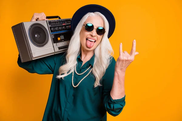 Photo of cool crazy babcia lady metal miłośnik muzyki magnetofon ramię pokazać rogi trzymać język usta nosić zielony koszula okulary naszyjnik retro czapka odizolowany żółty kolor tło — Zdjęcie stockowe