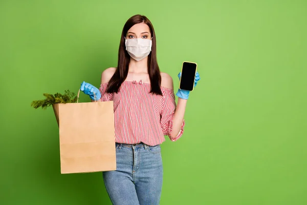 Dziewczyna shopper kupujący trzymać papier torba zakupy zioła smartfon polecam online dostawy nosić maskę medyczną niebieski lateks rękawiczki paski off-barki bluzka odizolowany zielony kolor tło — Zdjęcie stockowe