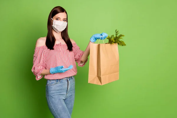 Fotoğrafçı kız medikal maske işçi kuryesi kağıt poşet teslimatı sebze sebze sebzeleri mavi lateks eldivenler kırmızı beyaz çizgili, omuzsuz bluz kot pantolon ve yeşil arka plan. — Stok fotoğraf
