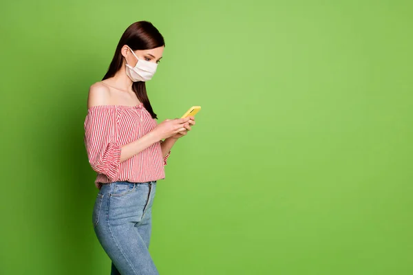 Side profil zdjęcie ładna młoda dziewczyna trzymać telefon czytać wiadomości numer zarażony świat nosić maskę dżinsy paski biały czerwony bluzka odkryte ramiona jasny zielony kolor tło — Zdjęcie stockowe