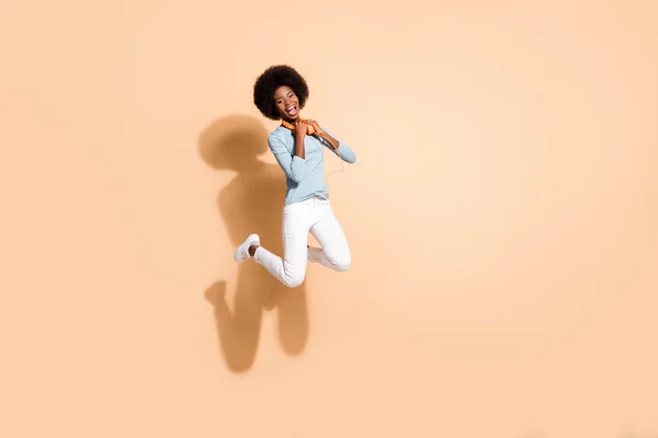 Foto retrato de menina americana africana alegre segurando fones de ouvido laranja vestindo em torno do pescoço pulando alto isolado no fundo de cor bege pastel — Fotografia de Stock