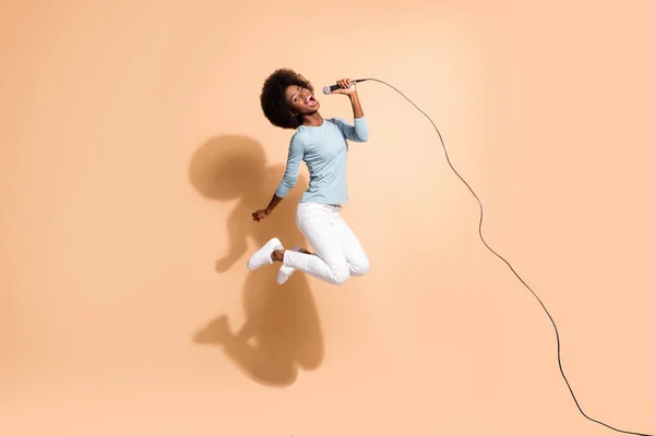 Foto retrato de chica afroamericana loca sosteniendo micrófono en una mano cantando en el aire aislado sobre fondo de color beige pastel — Foto de Stock