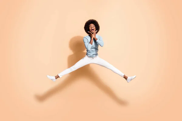 Foto retrato de mujer de piel negra sosteniendo dos manos cerca de la cara saltando con las piernas abiertas boca abierta aislada sobre fondo de color beige pastel —  Fotos de Stock