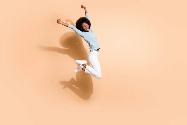 Фотопортрет черной кожаной девушки, прыгающей высоко вверх на два кулака в воздухе, приветствуя крики изолированных на пастельно-бежевом фоне — стоковое фото