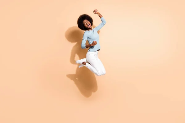 Фотопортрет брюнетки африканской женщины-американки, прыгающей вверх и празднующей изолированно на пастельно-бежевом фоне — стоковое фото