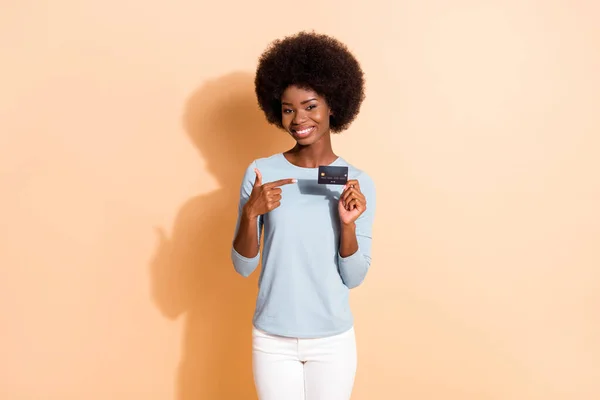 Retrato fotográfico de una chica sonriente de piel oscura apuntando con el dedo a una tarjeta de débito de plástico con un atuendo casual aislado sobre un fondo de color beige — Foto de Stock