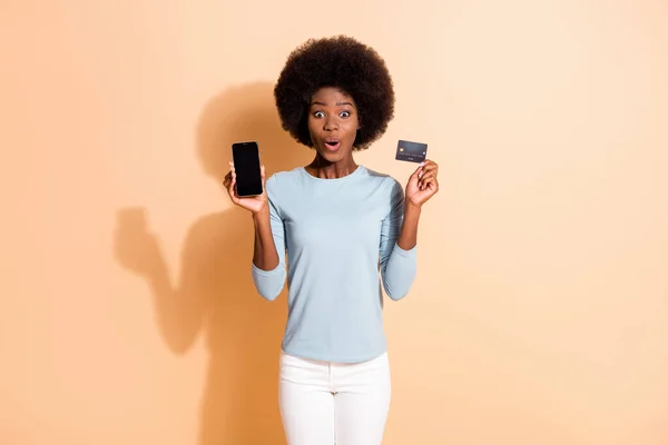 Foto retrato de escuro pele encaracolado menina chocado impressionado mostrando celular tela plástico cartão de crédito isolado no fundo cor bege — Fotografia de Stock