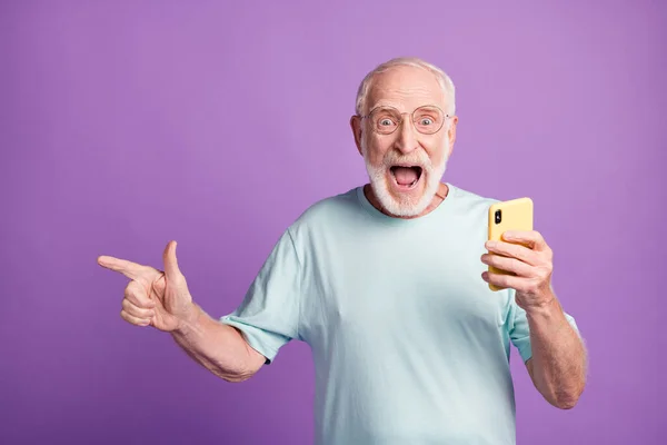 Photo portrait of excited man screaming pointing finger to side holding phone in one hand isolated on vivid purple colored background — Stock Photo, Image