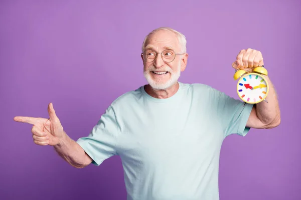 Retrato de hombre viejo divertido sosteniendo reloj que indica espacio vacío desgaste tela azul aislado sobre fondo de color púrpura —  Fotos de Stock