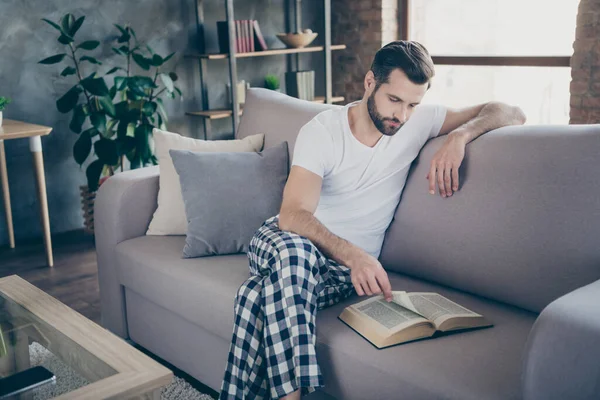 Retrato de su agradable chico barbudo enfocado atractivo sentado en diván leyendo interesante libro de papel página imprimir pasar tiempo libre ocio en el loft moderno estilo interior industrial en interiores — Foto de Stock