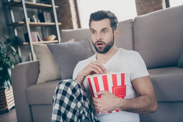 Foto de cara bonito chocado caseiro sentar confortável sofá chão quarentena ficar em casa assistir terror assustador filme tv comer pipoca boca aberta calças de pijama t-shirt sala de estar dentro de casa — Fotografia de Stock