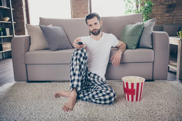 Full body photo of handsome guy homey sit comfy floor sofa quarantine stay home good mood watch movie tv eat popcorn hold remote controller pajama pants t-shirt living room indoors
