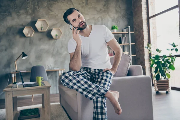 Photo of handsome man sit divan quantine home call friend smartphone tell talk talk talk style życia daily news wear checkered plaid pants white t-shirt in house in doors — Zdjęcie stockowe