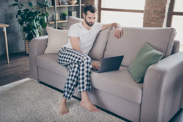 Retrato de su agradable chico morena enfocado atractivo sentado en el sofá trabajando en el ordenador portátil proyecto de TI puesta en marcha en el loft moderno apartamento interior industrial de la casa en interiores — Foto de Stock