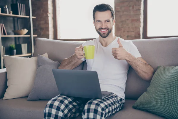 Retrato de su agradable atractivo alegre alegre morena chico sentado en el sofá dinking cacao trabajando remotamente mostrando thumbup en apartamento moderno loft casa interior industrial — Foto de Stock