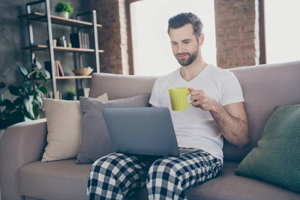 Foto de guapo homey chico sentado cómodo sofá bebida caliente café fresco navegación portátil freelancer trabajo remoto estancia casa cuarentena pijama fin de semana sala de estar en el interior — Foto de Stock