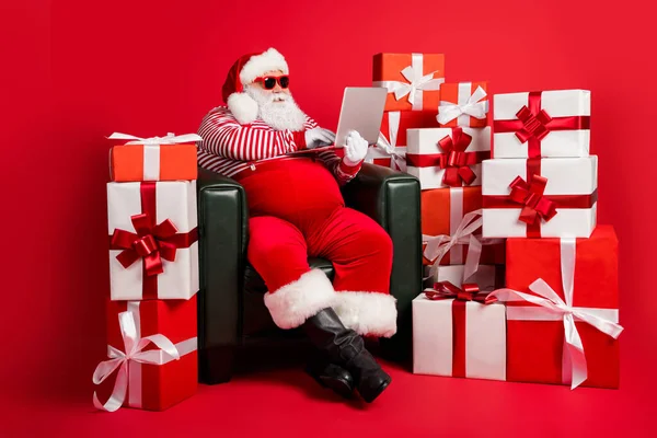 Noel Baba 'nın fotoğrafı koltuğa otur netbook kullan x-mas kostüm askısı tak gözlük izole edilmiş kırmızı renk arka plan — Stok fotoğraf