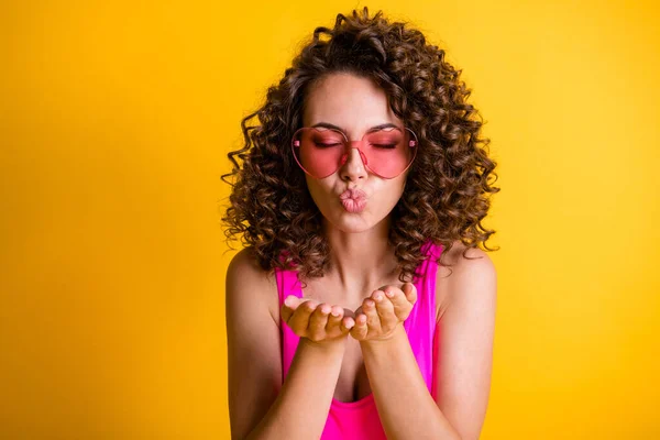 Foto van aantrekkelijke schattige meisjesachtige golvend kapsel dame blazen lucht kus open armen gesloten genieten oprechte gevoelens dragen roze hart vorm zonnebril tank top geïsoleerde gele kleur achtergrond — Stockfoto