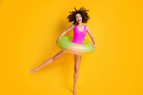 Bir bacağının üzerinde duran heyecanlı bir kadının fotoğrafı. Elinde yeşil şişme daire, iki eli çığlık atan pembe yüzme kıyafeti giyen canlı sarı arka planda izole edilmiş bir kadın. — Stok fotoğraf