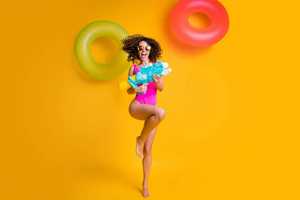 Foto retrato de alegre jovem morena de pé em uma perna segurando arma de água rindo vestindo fúcsia nadar desgaste isolado no fundo colorido amarelo vívido com anéis infláveis vermelhos verdes — Fotografia de Stock