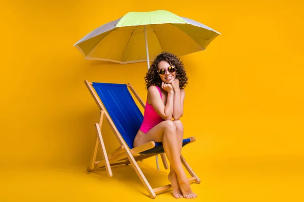 Şemsiyenin altında mavi şezlongda oturan sevimli bir kızın resmi. Elleri şemsiyeli, gözlüklü, pembe elbiseli, sarı renkli arka plan izole edilmiş. — Stok fotoğraf