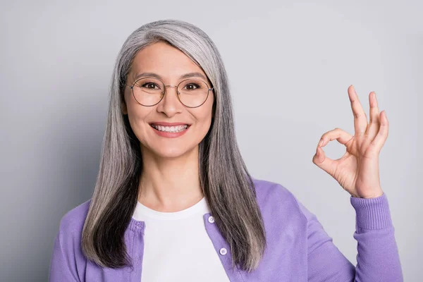 Foto de la señora pensionista encantadora pelo blanco levantar la mano mostrar okey toothy brillante sonrisa usar gafas camisa blanca cárdigan violeta aislado color gris fondo —  Fotos de Stock