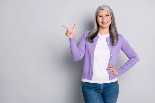 Foto portret van positieve optimistische oma lachend wijzend aan de zijkant met vinger in jeans violet vest bril geïsoleerd op grijze achtergrond — Stockfoto