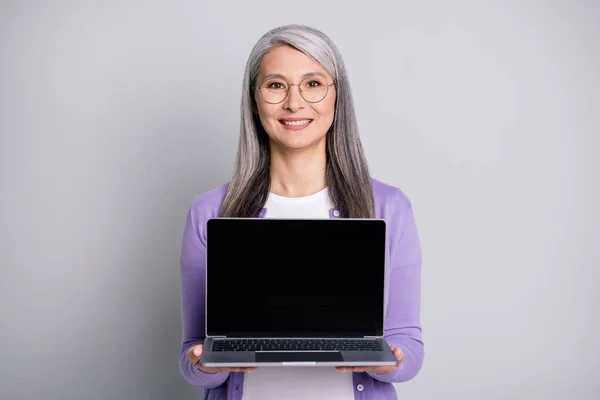 친절 한 나이든 여성이 웃고 있는 사진은 회색 배경에 고립 된 평범 한 보라색 가디건 안경을 착용하고 노트북을 열었다. — 스톡 사진