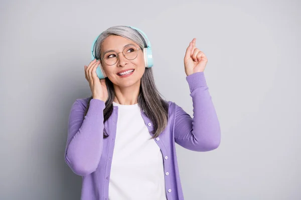 Retrato fotográfico de una mujer anciana sonriente soñadora mirando hacia arriba señalando con el dedo escuchando música tocando auriculares inalámbricos usando atuendo casual aislado sobre fondo de color gris —  Fotos de Stock