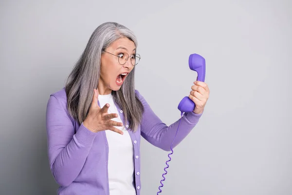 Фотографічний портрет старшої божевільної напруженої жінки, що кричить голосно тримаючи телефон з ретро-фіолетовим дротом ізольовано на сірому фоні — стокове фото