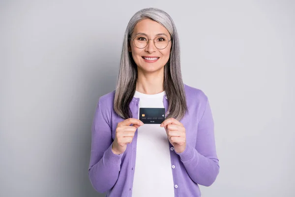 Foto ritratto di vecchia donna che tiene carta di credito con due mani che indossa cardigan viola isolato su sfondo grigio — Foto Stock