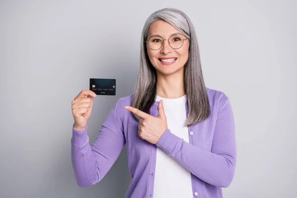 Foto retrato de mulher segurando cartão de banco de plástico apontando dedo para ele vestindo camisa violeta isolada no fundo de cor cinza — Fotografia de Stock