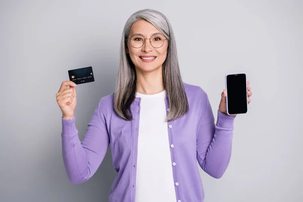 Foto ritratto di donna dai capelli bianchi grigi in possesso di carta di credito in plastica e smartphone con copyspace indossando casual cardigan viola isolato su sfondo grigio — Foto Stock