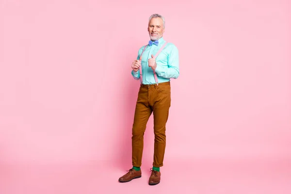 Retrato de cuerpo entero de caballero viejo alegre sonriente usando pantalones de corbata tocando tirantes aislados sobre fondo de color rosa —  Fotos de Stock