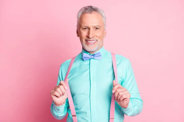 Foto porträtt av snygg attraktiv senior man med toothy leende hålla hängslen bowtie skjorta isolerad på rosa färg bakgrund — Stockfoto