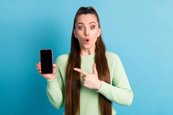 Фотографічний портрет шокованої дівчини, що вказує пальцем по телефону з порожнім простором у зеленому джемпері ізольовано на пастельному синьому фоні — стокове фото