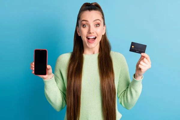 Retrato fotográfico de una chica excitada con la boca abierta sosteniendo el teléfono con tarjeta bancaria en blanco usando un jersey verde aislado sobre fondo de color azul pastel —  Fotos de Stock