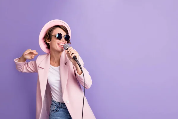 Foto de linda encantadora morena cabello joven dama usar gorra abrigo gafas cantando en micrófono aislado sobre fondo de color violeta —  Fotos de Stock