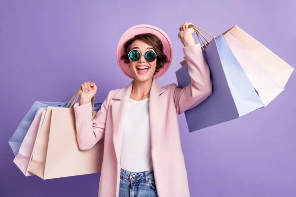 아주 긍정적 인 갈색 머리 여자가 쇼핑하는 것을 보고 놀라는 사진은 보라색 배경 위에 분리 된 안경 핑크 모자를 쓰고 있다 — 스톡 사진
