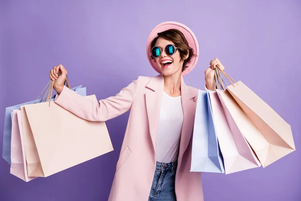 Φωτογραφία από γλυκό αρκετά αισιόδοξη μελαχρινή νεαρή κοπέλα μεταφέρουν τσάντες κατάστημα φορούν γυαλιά ροζ παλτό καπάκι απομονώνονται σε μωβ φόντο χρώμα — Φωτογραφία Αρχείου