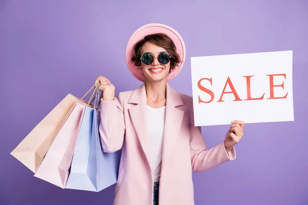 Retrato de optimista dulce dama mantenga compras venta banner usar gafas capa gorra rosa aislado sobre fondo púrpura —  Fotos de Stock