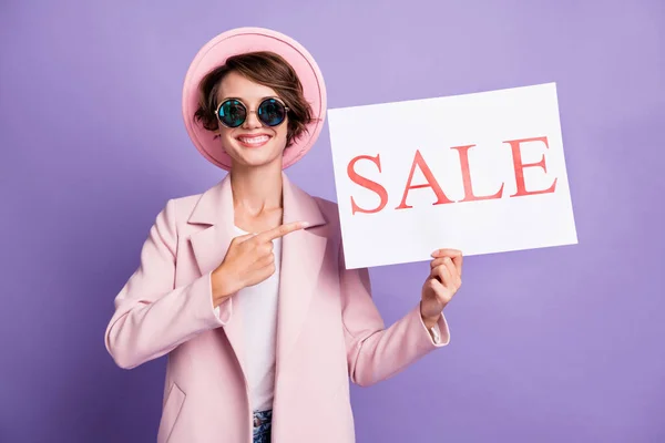Foto von ziemlich fröhlich junge Dame hält in der Hand Verkauf promo tragen Mantelmütze Brille isoliert auf pastelllila Hintergrund — Stockfoto